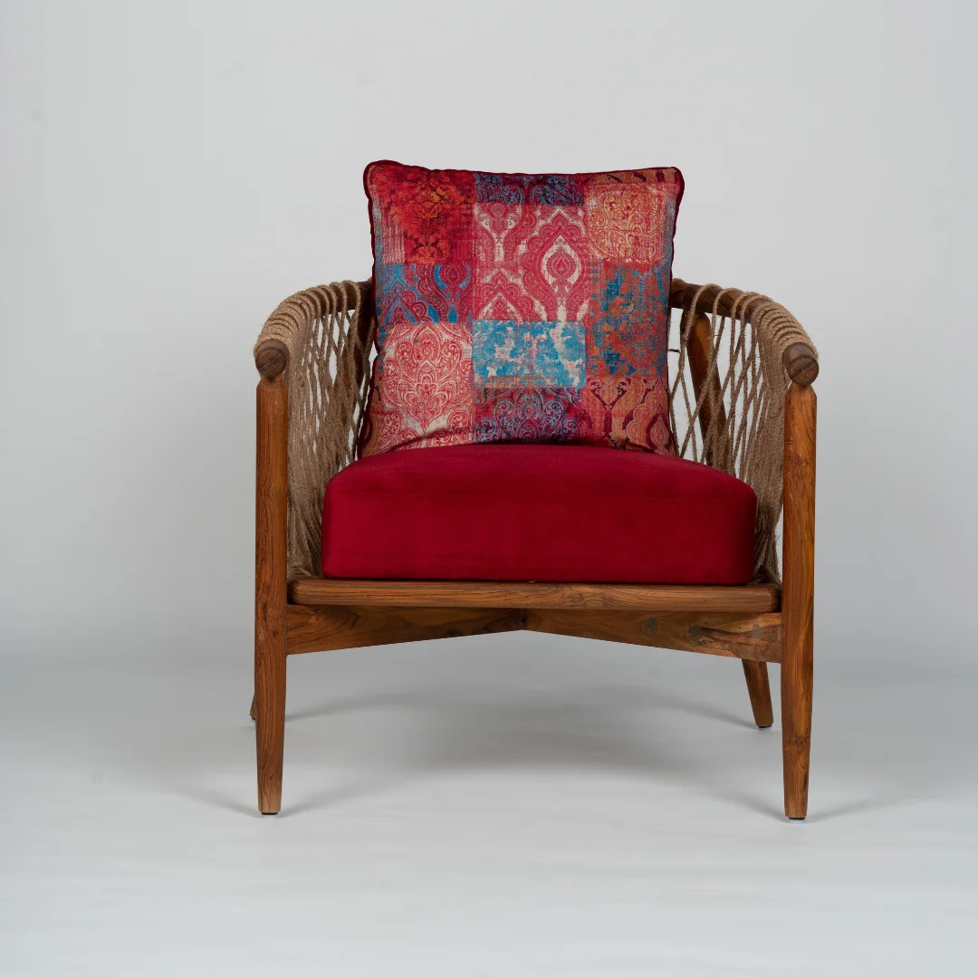 A stylish reclaimed wood accent chair featuring a sturdy wooden frame with woven rope details on the sides. The chair is complemented by a rich red cushion and a colorful patterned pillow, adding warmth and personality to the space. This piece combines natural wood elements with bold, vibrant textiles, making it a perfect accent for any room. The unique design balances both comfort and aesthetic appeal, ideal for adding character to interiors.
